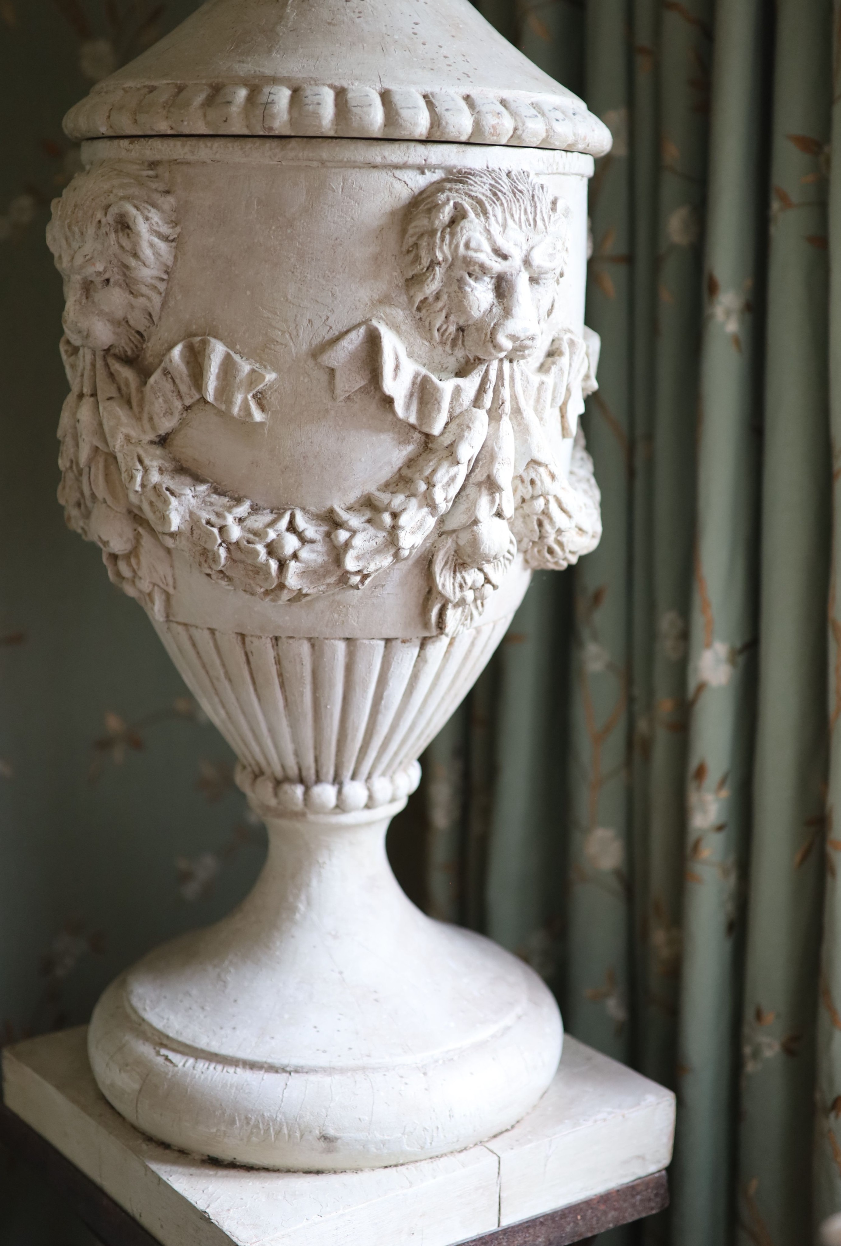 A pair of white painted carved walnut urns and covers, on wrought iron stands, the urns carved with fruit, lions masks and floral swags, height 90cm, the tapered stands with lattice work sides, height 90cm, overall heigh